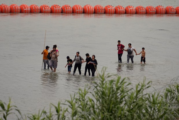 Migrants who crossed the Rio Grande from Mexico walk past large buoys being deployed as a border barrier on the river in Eagle Pass, Texas, Wednesday, July 12, 2023. The floating barrier is being deployed in an effort to block migrants from entering Texas from Mexico.